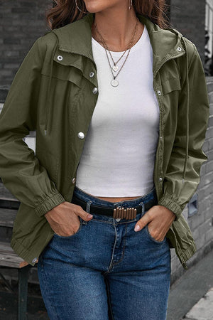 Woman wearing olive green outdoor hooded raincoat with elastic cuffs and snap-button closure over white top and jeans