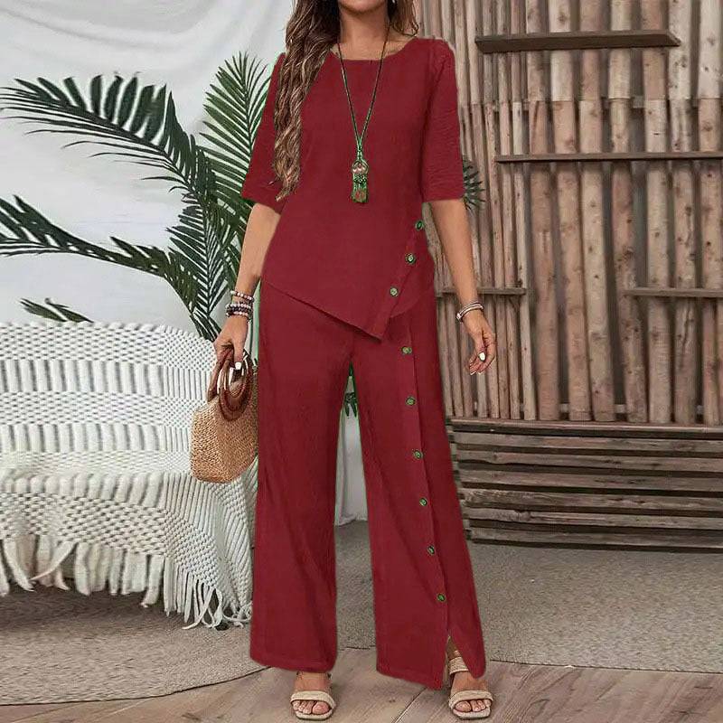 Woman wearing rust red Asymmetry Flow Co-ord with button-front top and flowing pants, standing indoors by bamboo wall and plants