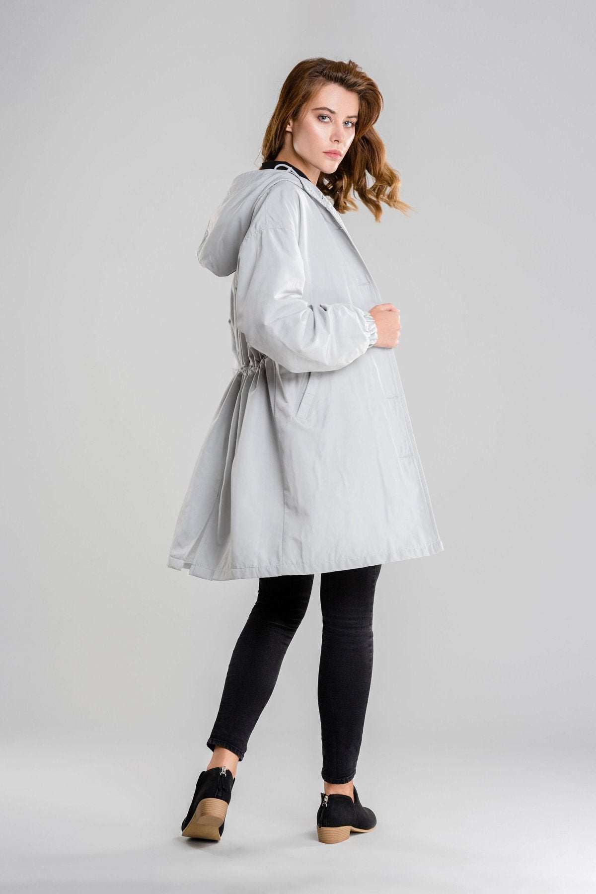 Woman wearing light gray water resistant oversized hooded windbreaker rain jacket with black leggings and shoes standing sideways on gray background