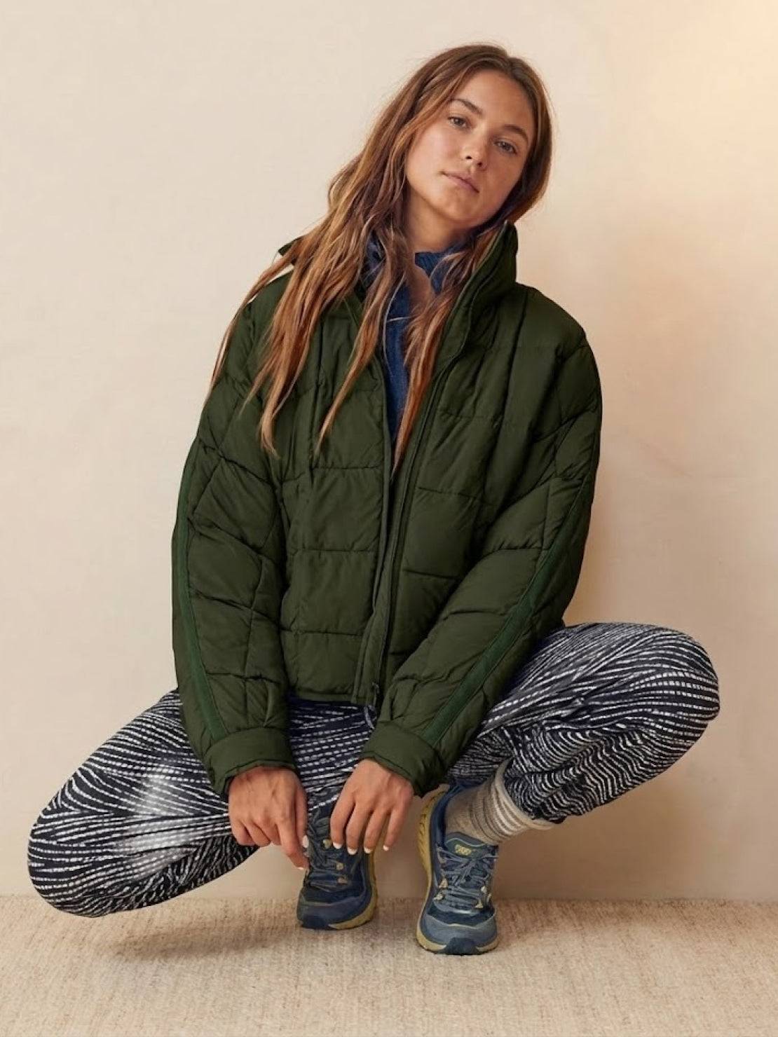 Woman wearing lightweight green puffer travel jacket with patterned leggings and sneakers, sitting in a crouched pose against a neutral background.