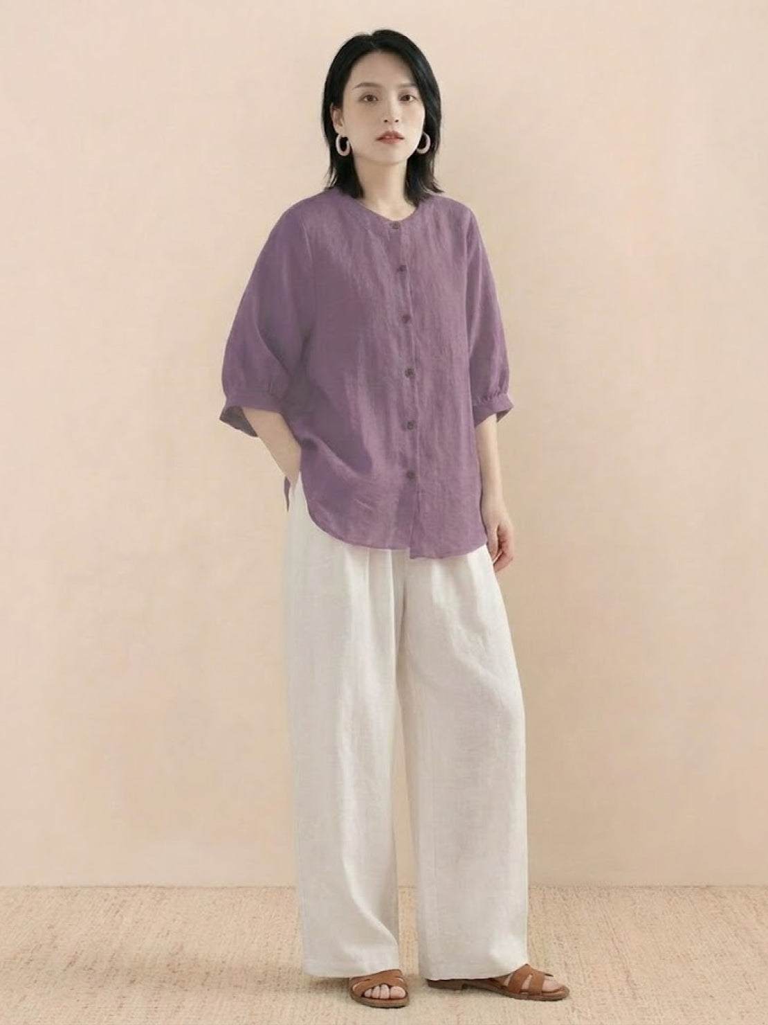 Woman wearing relaxed loose breathable linen purple blouse and white wide-leg pants standing against neutral background
