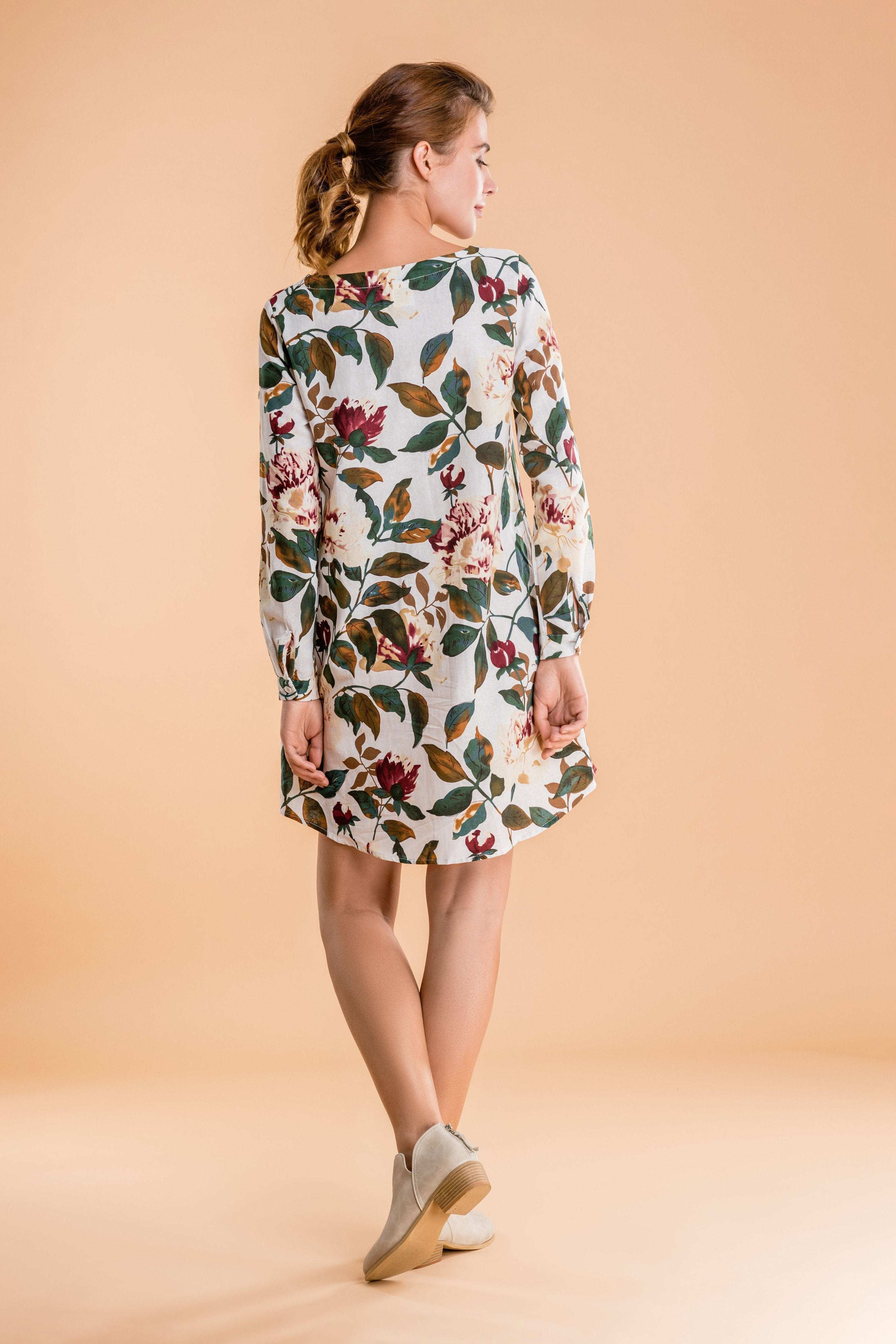 Woman wearing an all over floral print linen shift dress with long sleeves and beige shoes against a peach background.