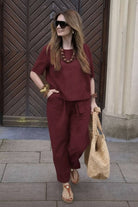 Woman wearing a relaxed fit burgundy cotton linen set with short-sleeve top and straight-leg pants, styled with sunglasses and a woven tote bag.