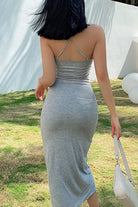 Back view of woman wearing elegant side slit midi dress in grey with crisscross straps, holding white handbag outdoors.