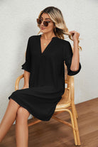 Woman wearing a black Dainty Charm Casual Ruffle Dress with ruffled V-neck and short sleeves sitting on a wooden chair indoors
