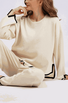Woman wearing a beige long sleeved relaxed fit lounge two-piece set with black trim accents sitting on the floor.