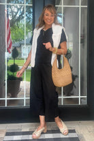 Woman wearing black loose fit co-ord set with a white knit vest, carrying a woven bag and standing in front of glass doors
