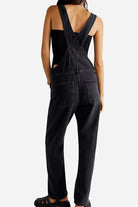 Back view of woman wearing black denim-look tapered-leg adjustable bib overalls with cross-back straps and pockets