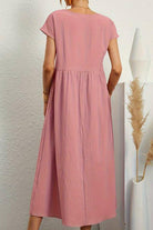 Back view of a pink capped-sleeve voluminous pleated midi dress on a woman against a white textured wall and decorative vases.