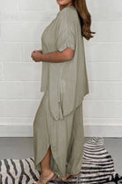 Woman wearing relaxed linen-like travel 2-piece set in beige with loose fit and side slits, standing indoors against white brick wall.
