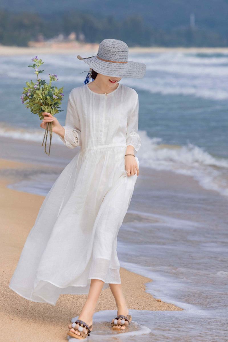 woman wearing white 100 linen coastal shift dress and sun hat walking on the beach holding flowers