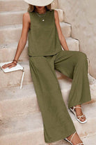 Woman wearing olive green sleeveless wide leg pants set sitting on stairs with white sandals and hat