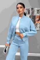 Woman wearing The Voyager Zip Set in light blue, consisting of a zip-front sweatshirt and wide-leg joggers, styled with a white crop top and silver clutch.