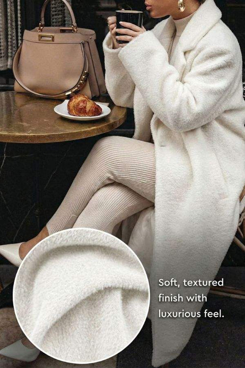 Woman wearing white oversized teddy coat with plush texture sitting at cafe table with handbag and croissant