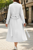 Woman wearing a timeless retro belted trench coat in white, showing back view with full-length cut and waist belt on city street.