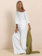 Woman wearing a white short-sleeved pullover and wide trouser matching set holding a green jacket.