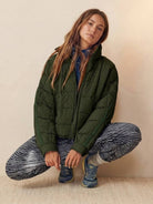 Woman wearing lightweight green puffer travel jacket with patterned leggings and sneakers, sitting in a crouched pose against a neutral background.