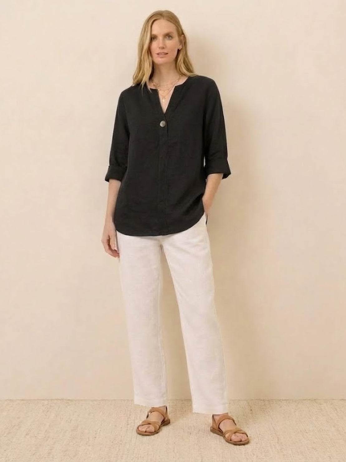 Woman wearing black pure linen age-defying shirt with button detail and white pants, standing against beige wall