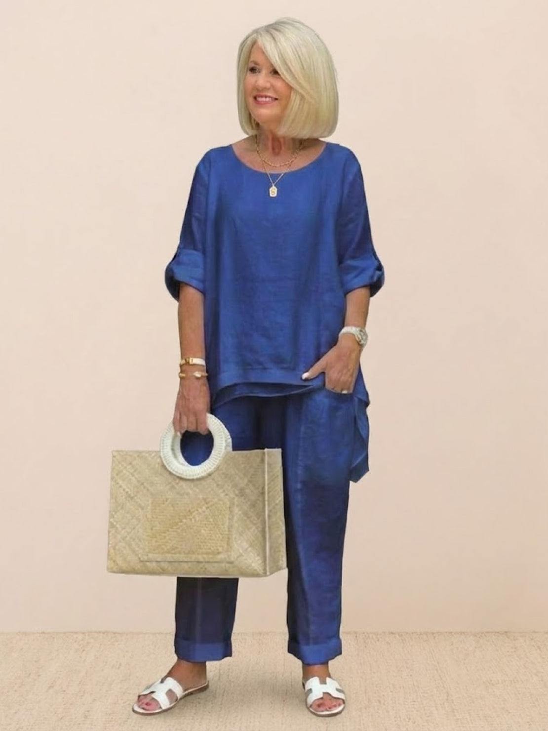 Woman wearing blue asymmetrical two-piece trouser set with rolled sleeves and holding a beige handbag.