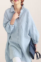 Light blue oversized linen shirt with relaxed fit, button front, and rolled sleeves worn by a woman holding a bag.