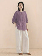 Woman wearing relaxed loose breathable linen purple blouse and white wide-leg pants standing against neutral background