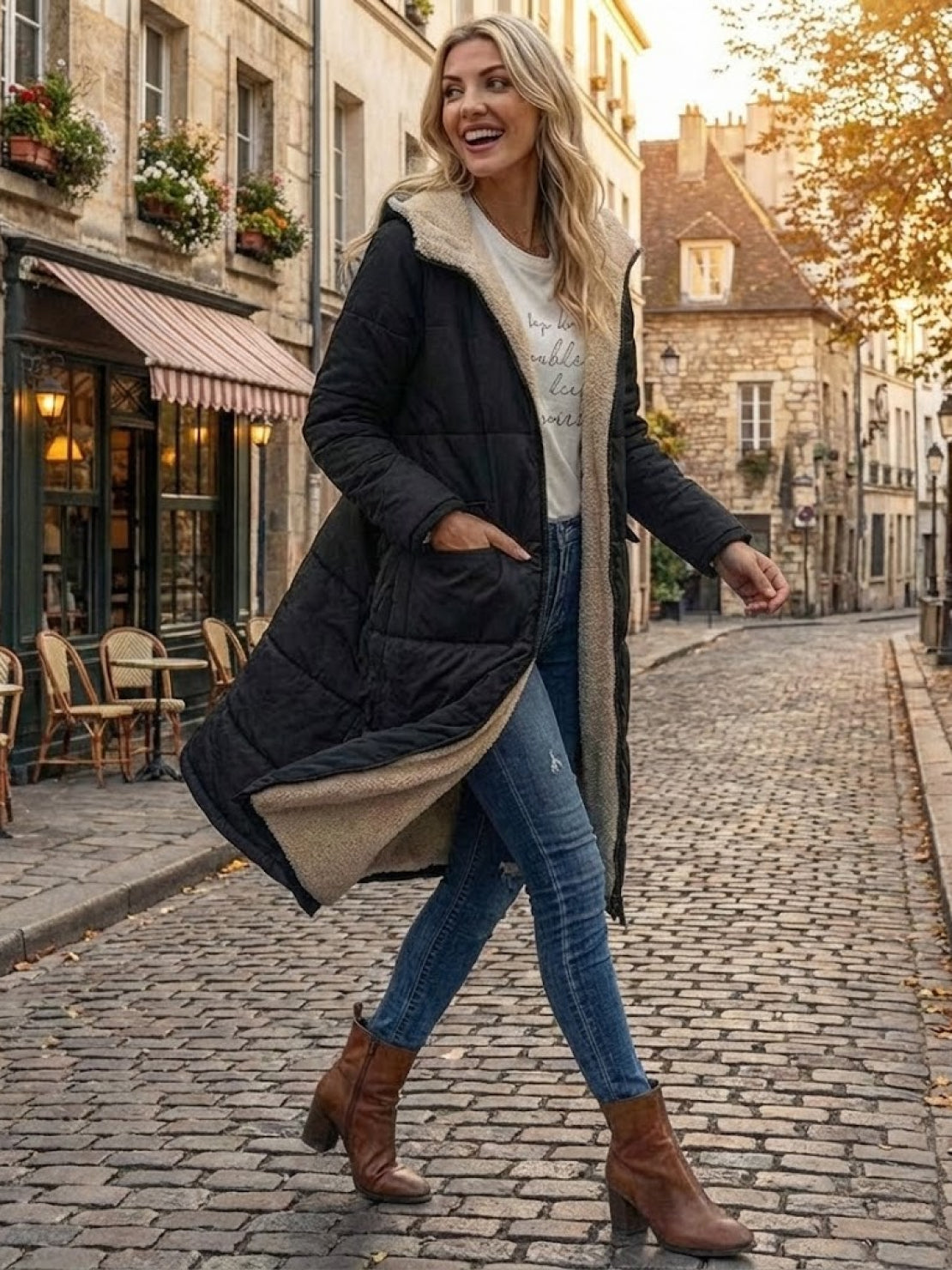 Woman wearing black sherpa-lined hooded teddy coat with jeans and brown boots walking on cobblestone street in autumn background