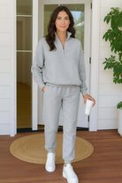 Woman wearing light gray cotton blend tracksuit with zip neckline and cropped hem, standing on wooden porch with white sneakers and holding a coffee cup