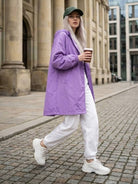 Woman wearing oversized purple Rain Couture Windbreaker with white pants, white sneakers, and green cap walking outdoors with coffee cup