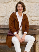 Woman wearing brown Rain Couture Windbreaker with hood, white shirt, and white pants sitting on wooden bench outdoors