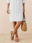 woman wearing knee-length white cotton dress with 3/4 sleeves holding woven straw bag and brown sandals