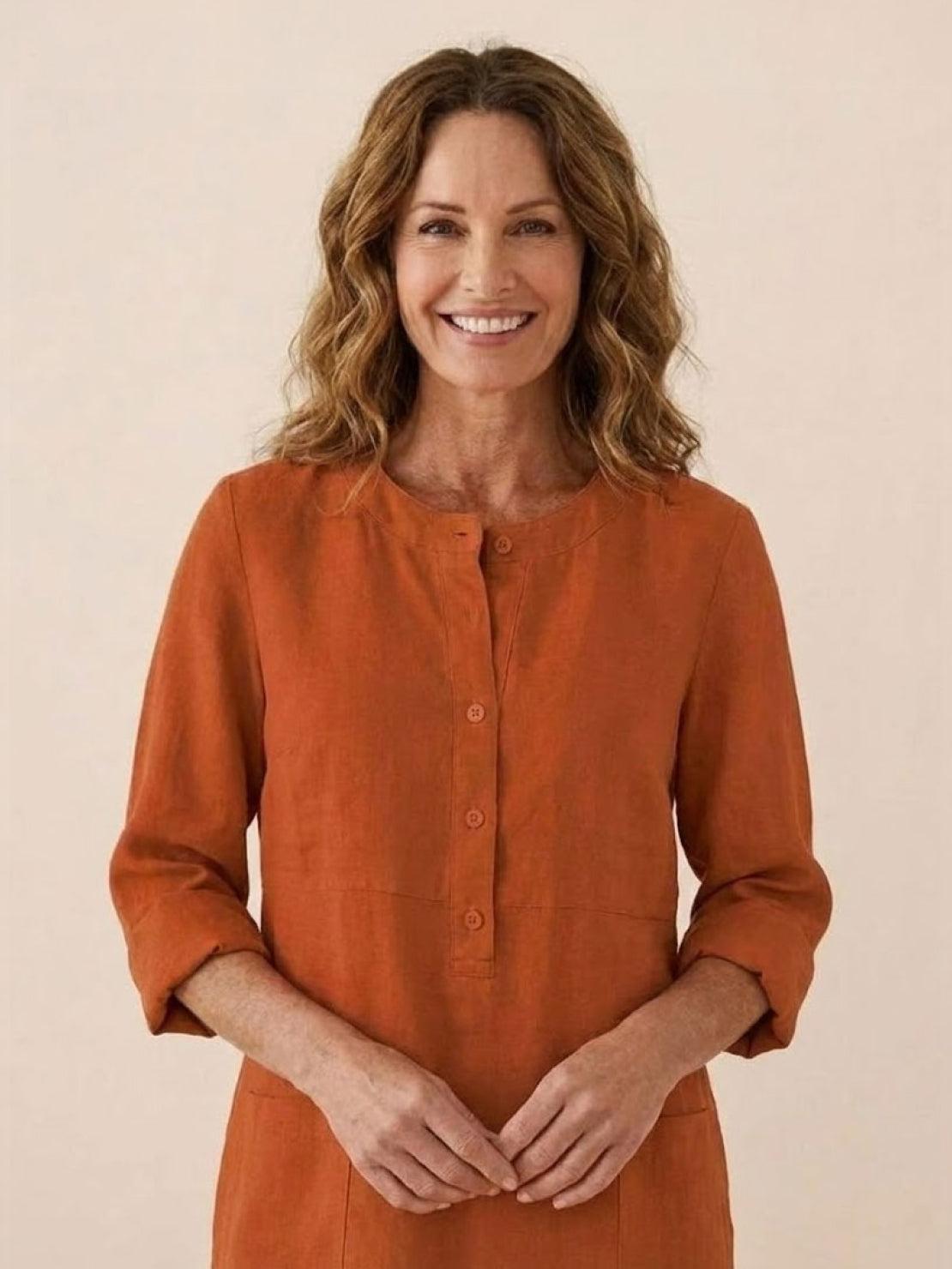 Woman wearing a rust-colored cotton dress with rolled ¾ sleeves and button front detail smiling against a plain background