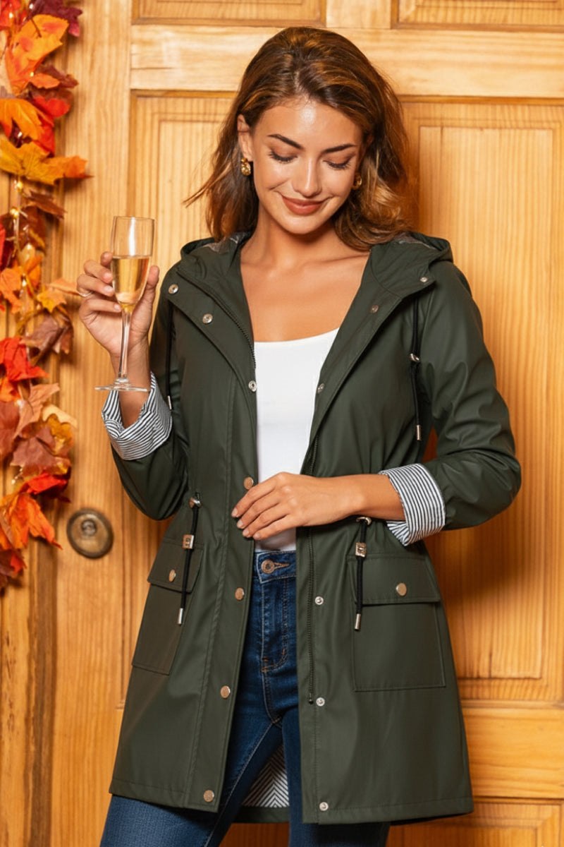 Woman wearing green water resistant windbreaker jacket with hood and striped lining, holding a glass of champagne indoors